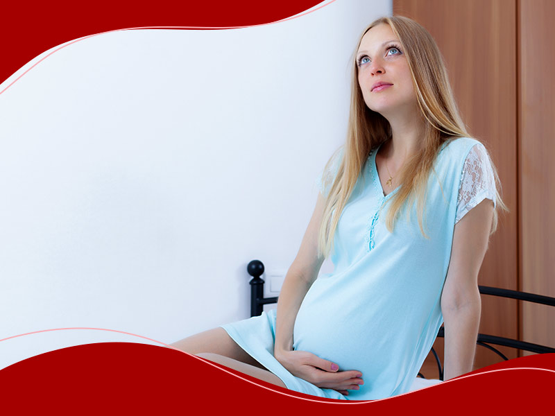 Mulher grávida, sentada na cama de vestido azul, segurando a barriga e olhando para cima, pensando se o melhor é colocar a prótese de silicone antes ou depois da gravidez.