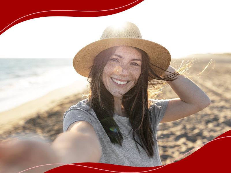 Mulher de camiseta cinza e chapéu tirando selfie feliz na praia